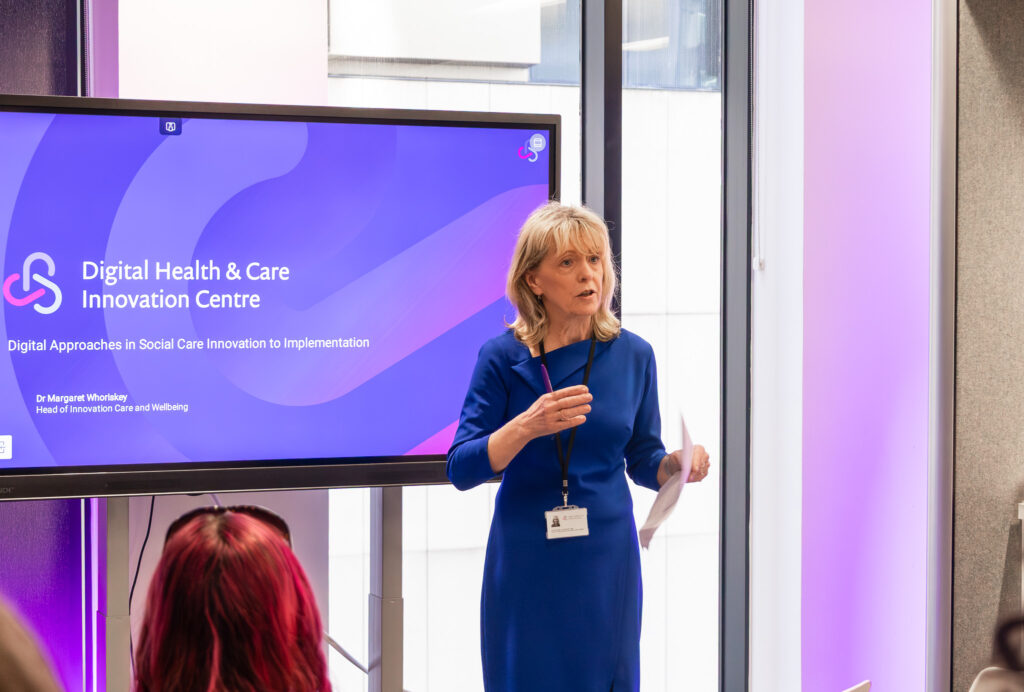 Dr Margaret Whoriskey, Head of Innovation for Care and Wellbeing at the Digital Health Care Innovation Centre DHI, presenting on Digital Approaches in Social Care Innovation to Implementation at a DHI event, standing in front of a branded screen displaying the DHI logo and presentation title