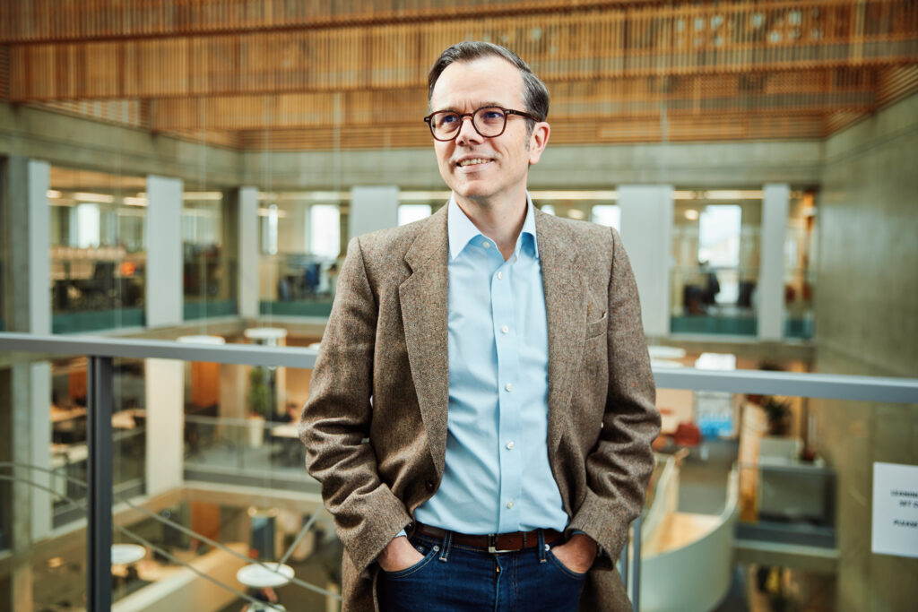 Professor Michael Rovatsos, Chair of Artificial Intelligence and Dean of Research and Innovation in the College of Science and Engineering at the University of Edinburgh, photographed in a modern open-plan university building with glass balustrades and a wooden ceiling, speaking to the University of Edinburgh's role in AI governance and the Edinburgh Data and AI Exchange report Governing the Future