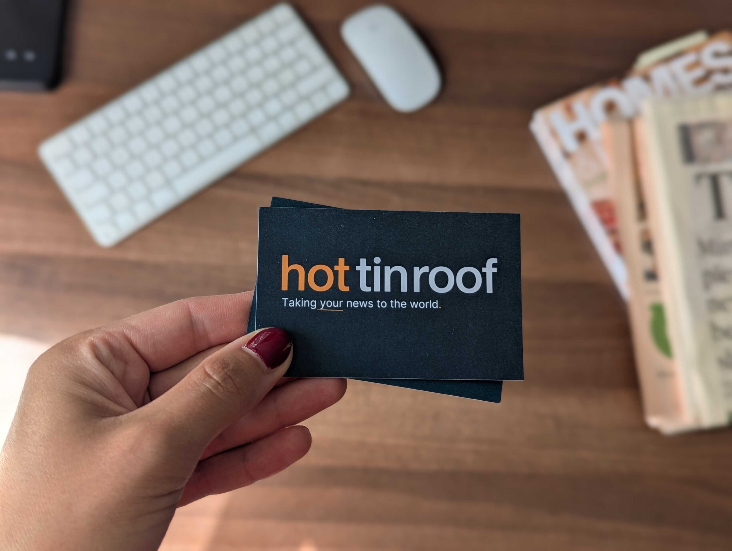 hot tin roof business card being held up over a desk with keyboard and newspapers