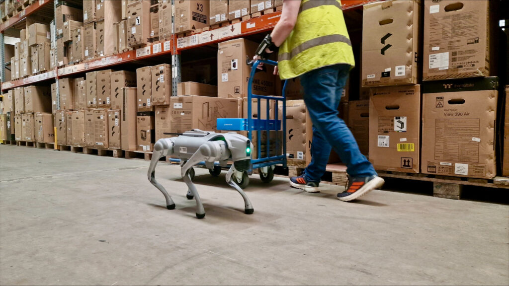 Unitree quadruped robot walking beside a warehouse worker pushing a trolley between shelves of boxed computer components, image via Hot Tin Roof PR agency