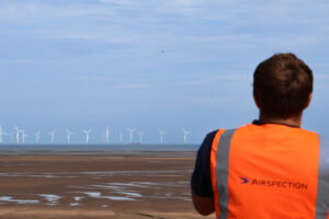 Airspection engineer in an orange hi-vis vest watches a drone fly over Liverpool Bay towards an offshore wind farm – Hot Tin Roof PR agency