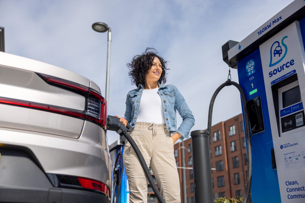 Smiling woman in a denim jacket charges a silver electric car at a Source ultra-rapid EV charger – Hot Tin Roof PR agency