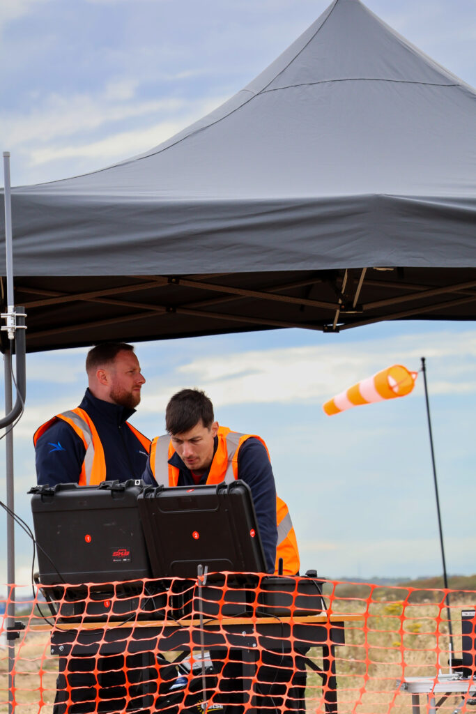 Two Airspection engineers in hi-vis vests monitor a drone flight from a ground control station beside a windsock – Hot Tin Roof PR agency