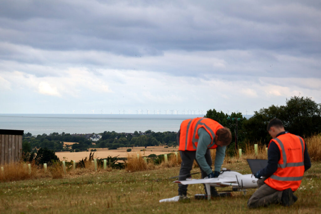 Two Airspection engineers prepare a white long-range drone on a coastal hillside with an offshore wind farm on the horizon – Hot Tin Roof PR agency
